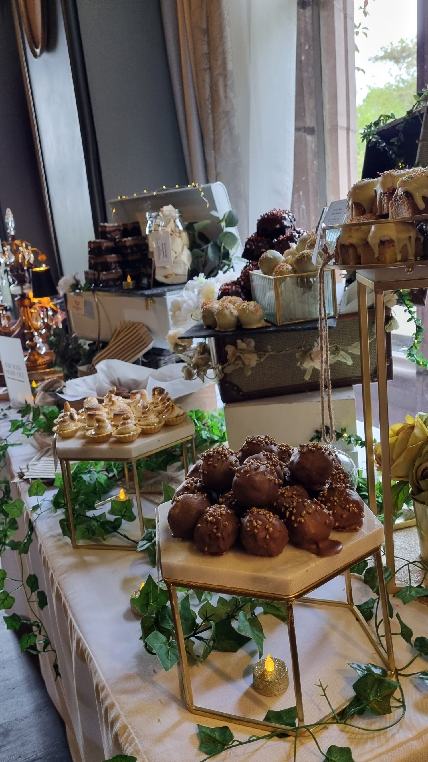 Dessert table with various treats and decorations, including a gold hexagonal stand and greenery. Set up at wedding venue Ellingham Hall in Northumberland, the north east.