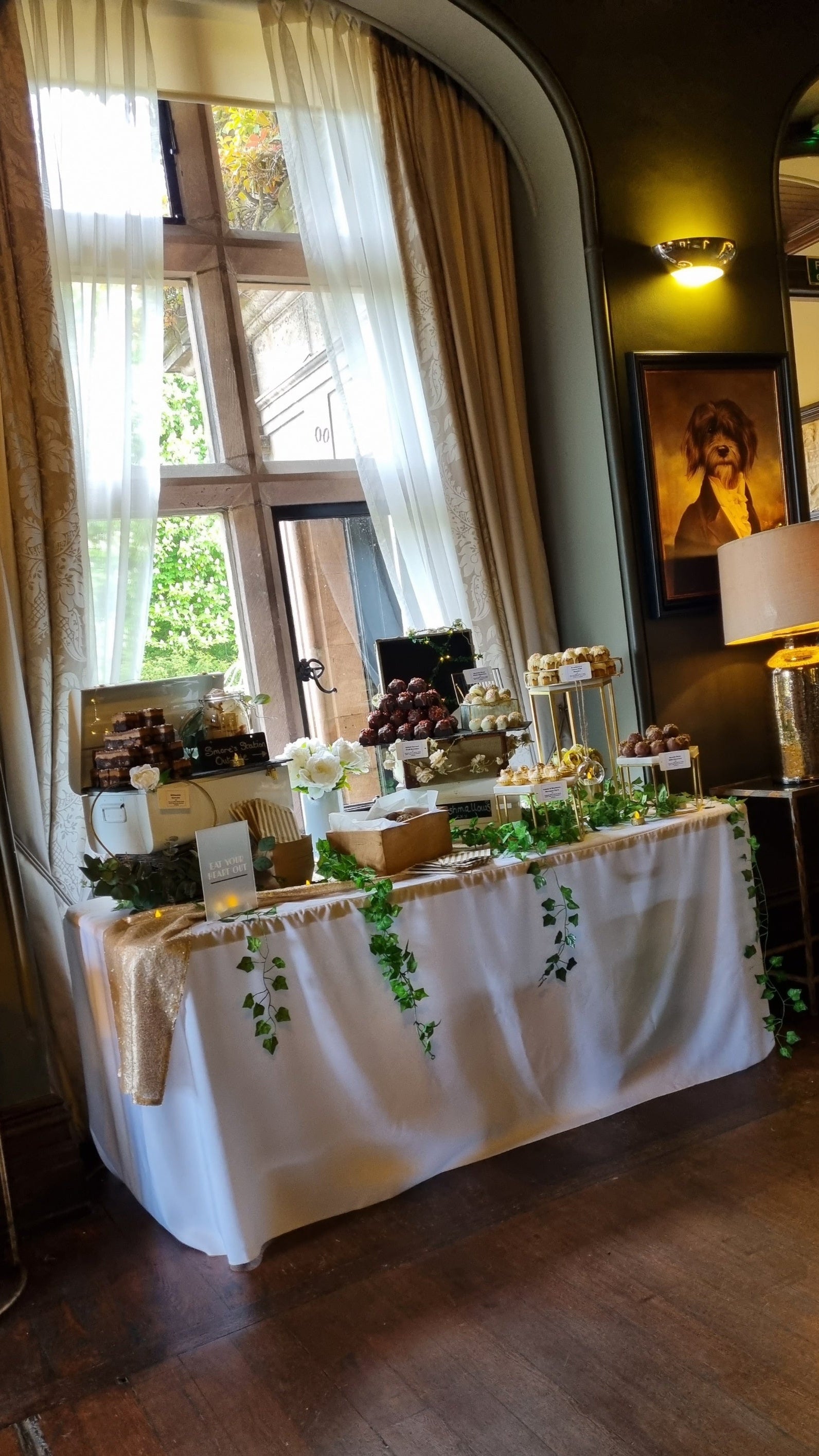 Decorated dessert table with greenery in a room with large windows and a lamp at Ellingham Hall Northumberland in the North east. Table is filled with layers of sweet treats, heaped upon boxes and slates.