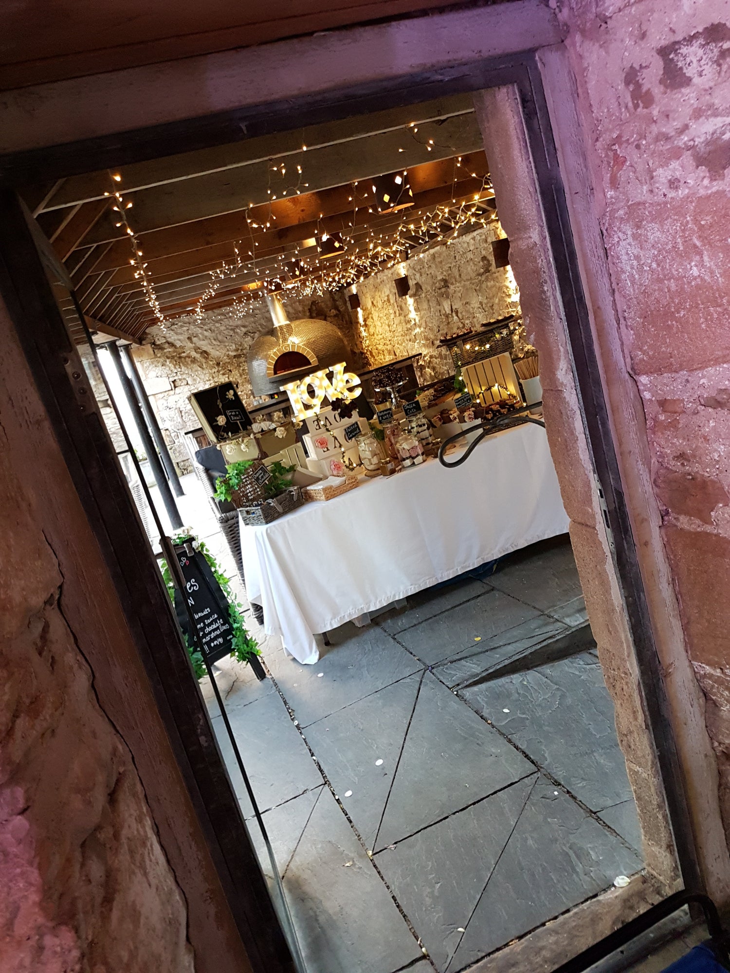 View of a barn door open at a wedding venue, showcasing an exterior courtyard through a doorway. In that view is a dessert table laid with display boxes and piled high with desserts and marshmallow jars.