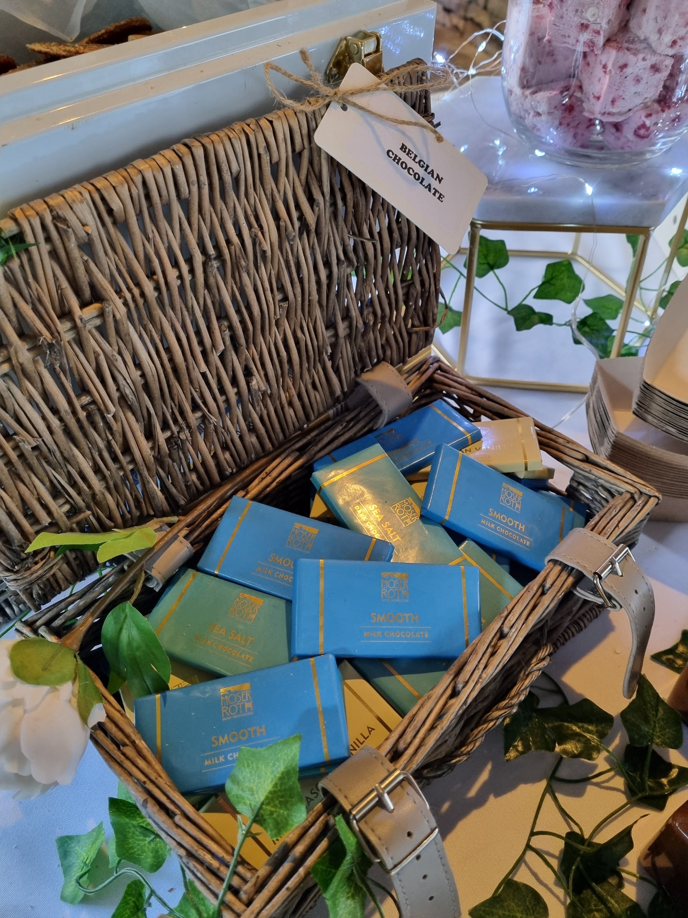 Basket with blue and gold chocolate packages inside, surrounded by greenery and a white surface.