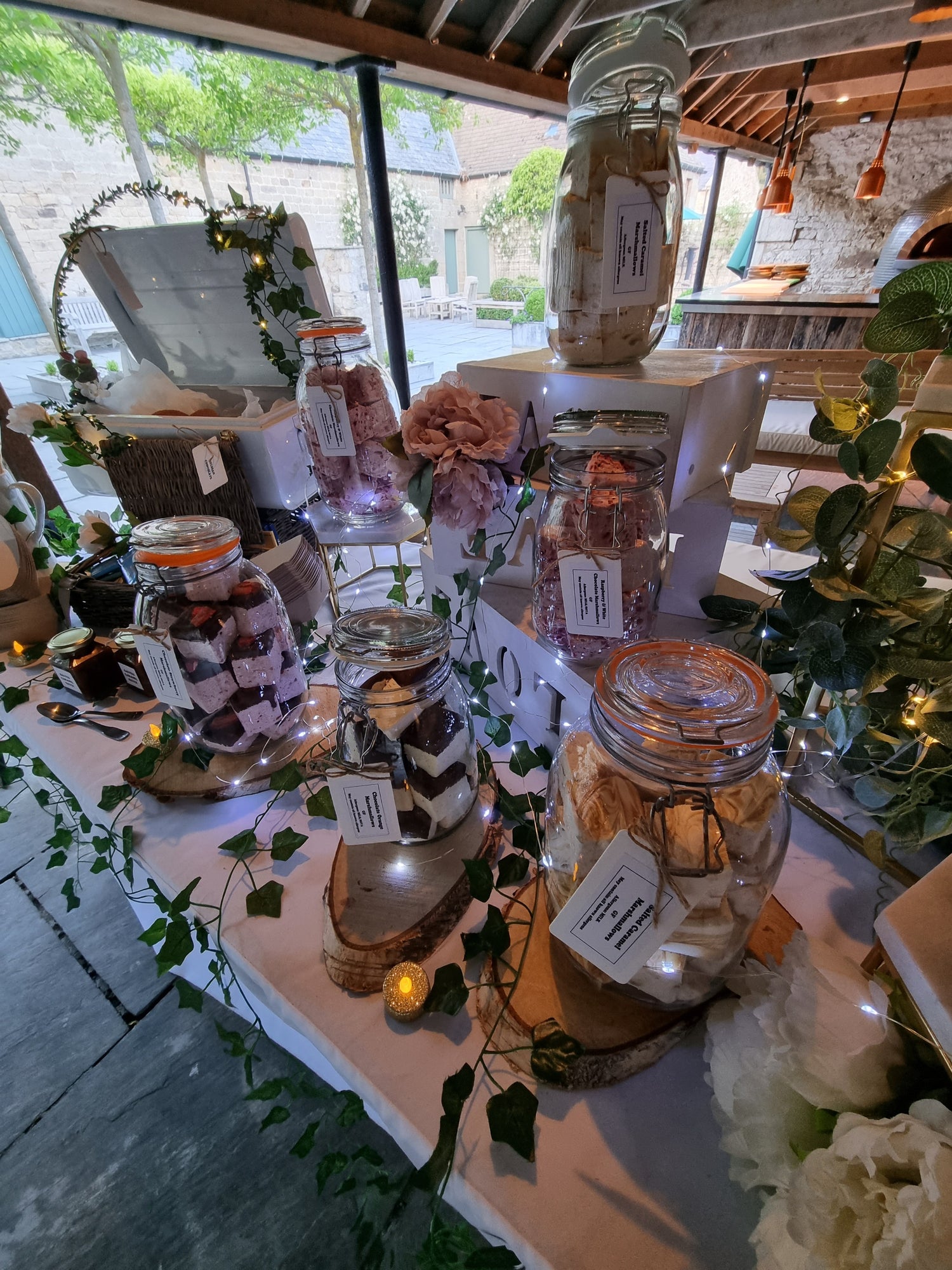 Full set up of a wedding smores station set up at Healey Barn wedding venue in Northumberland North East England. Scene includes green foliage, layered & stacked boxes with log cuts that hold glass jars filled with handmade marshmallows, salted caramel, biscuits and chocolate bars.
