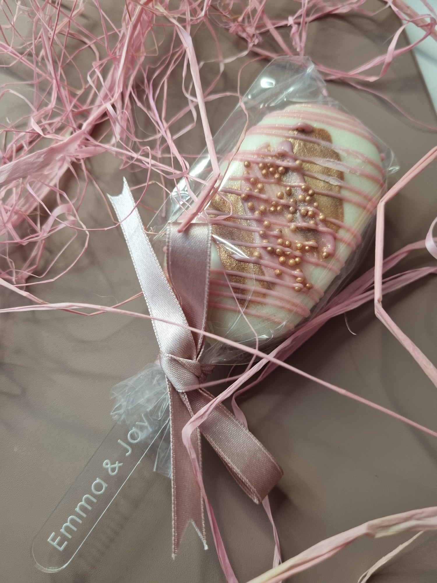 Decorative chocolate dipped marshmallow lolly item wrapped in pink ribbon on a textured surface with 'Emma & Jay' clear lolly stick