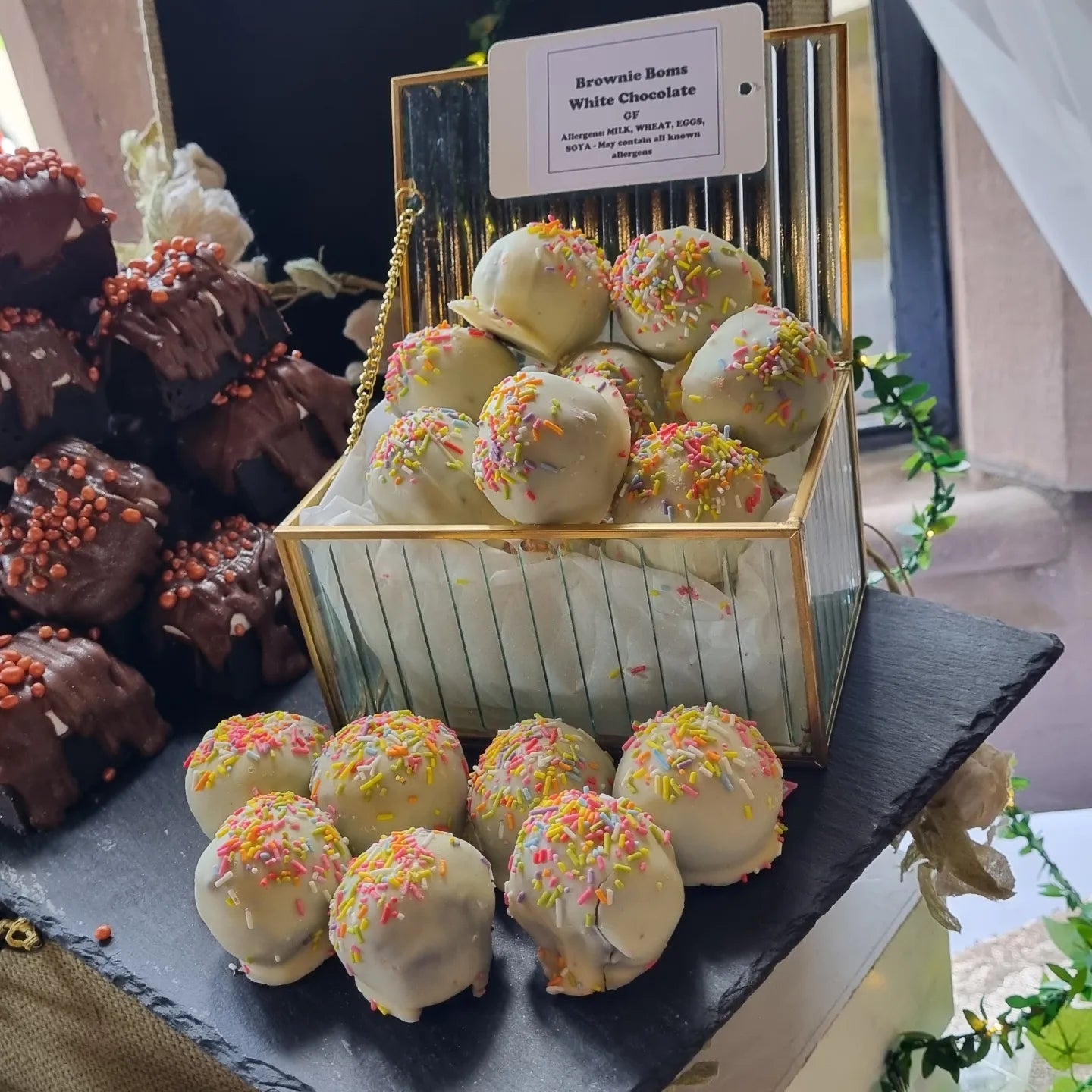 Decorative box of white chocolate brownie truffles with colorful sprinkles on a black slate surface. Brownies in the background on s dessert table at Ellingham Hall wedding venue