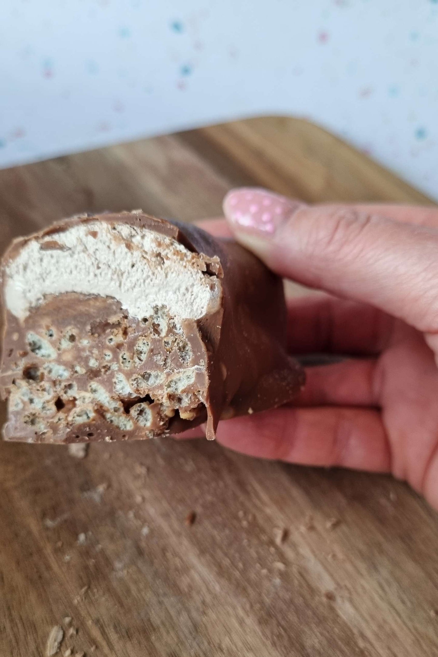 Handmade Chocolate Bar - Double Decker held by hand. layered chocolate crispies with a nougat and caramel top then all dipped in chocolate. The background is a wooden board on top of a table.
