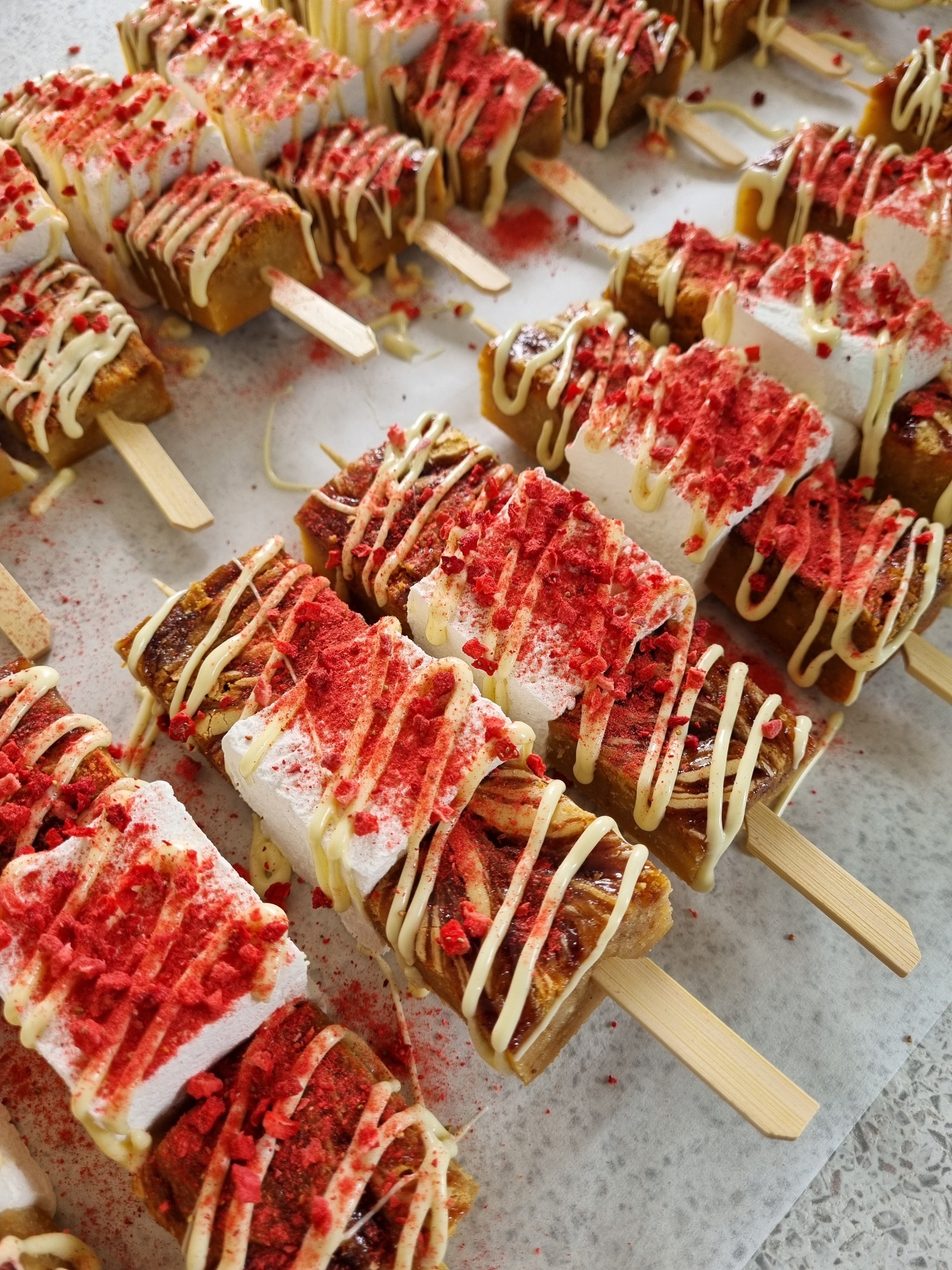 Dessert blondie & marshmallow skewers with raspberry bits and white chocolate toppings on a light surface
