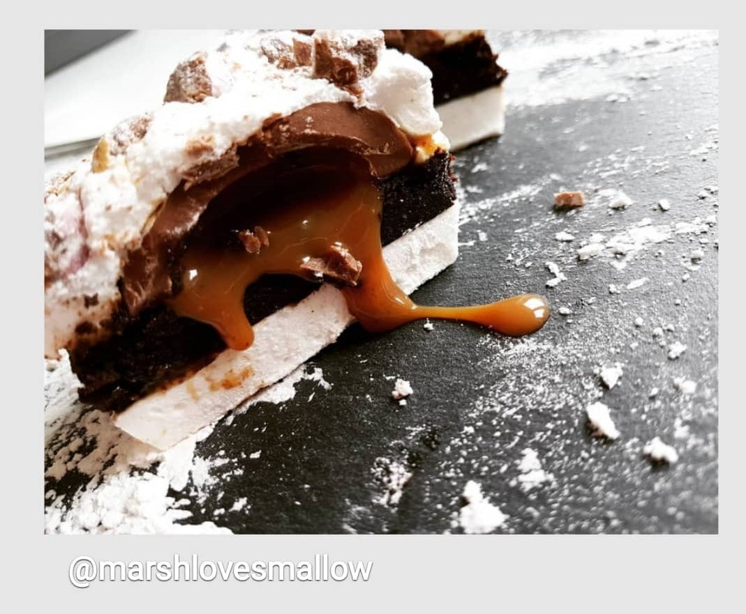 a brownie sandwiched between marshmallow, inside the brownie is a gooey caramel egg that is oozing caramel sauce. It is placedon a grey table with a dusting of white icing sugar.