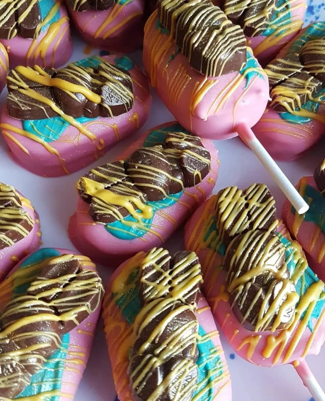 A mound of lollies on a white table. The marshmallow pops are pink and have been painted with bluey green chocolate, drizzled with yelloe chocolate and finished with a chocolate easter bunny.