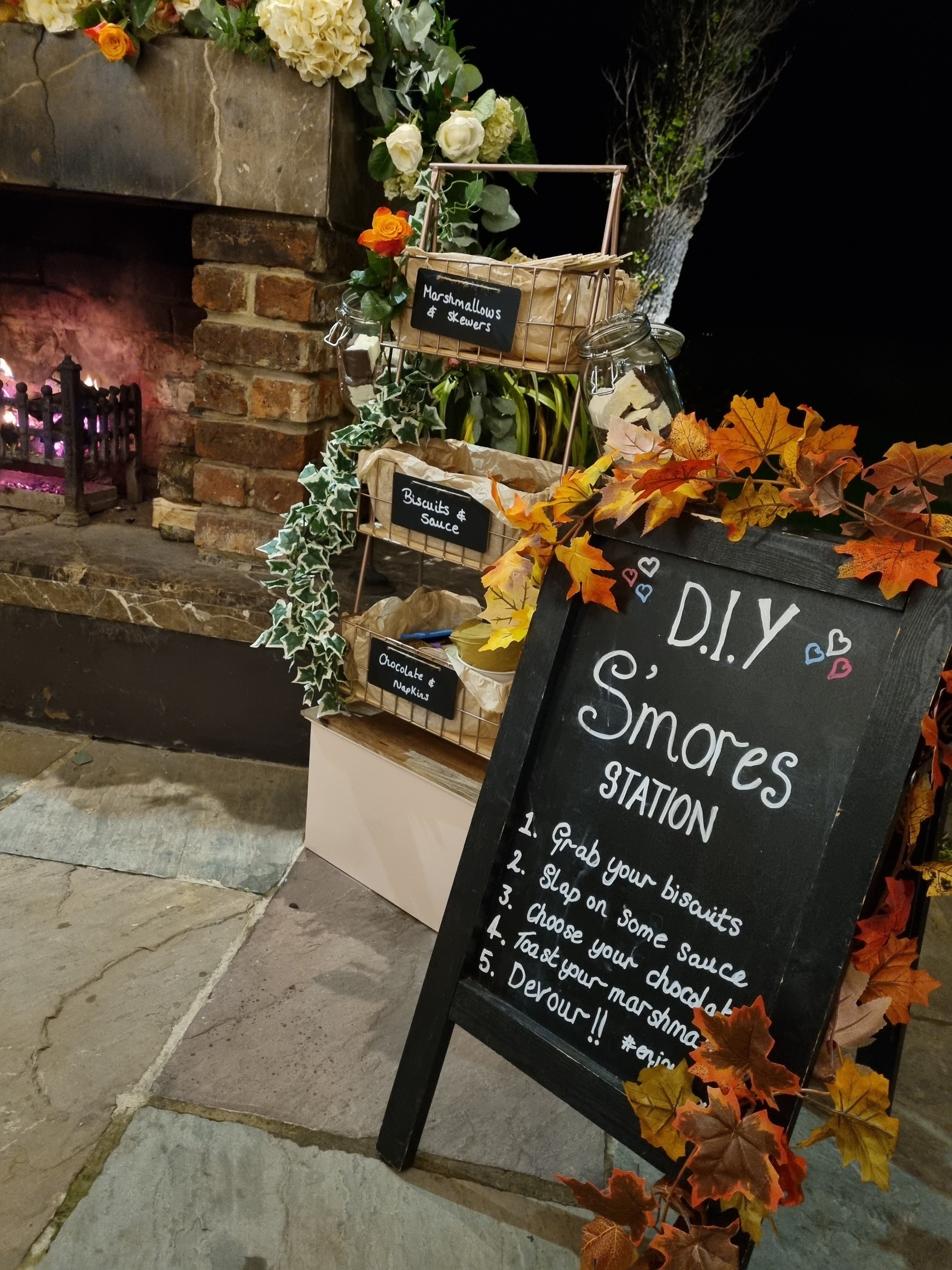 Outdoor setting at Newton Hall Northumberland north east, with a DIY S'mores station sign, baskets, and decorative elements near a fireplace.