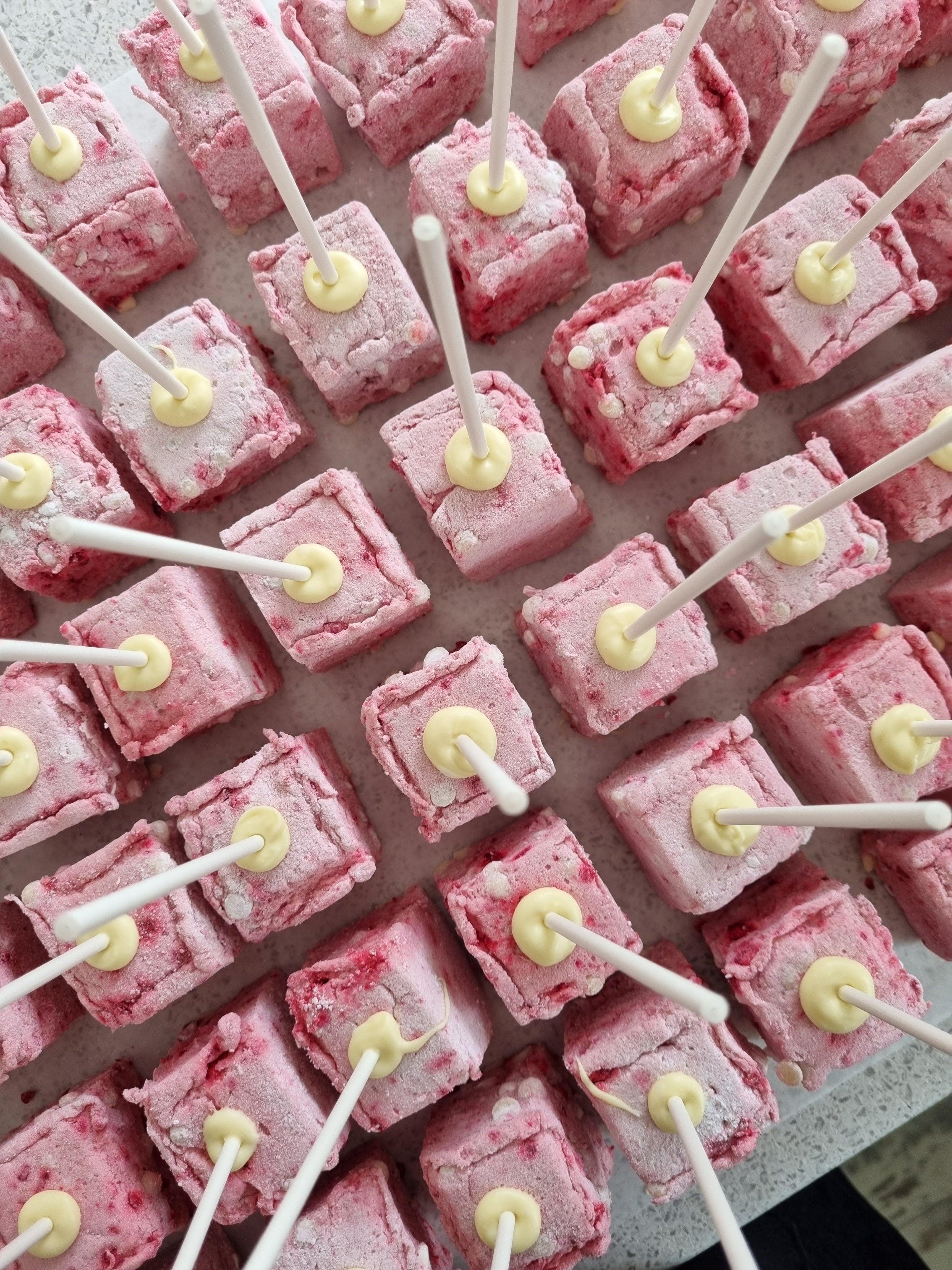 Pink square marshmallow treats on white sticks arranged in a grid pattern on top of table looking from above