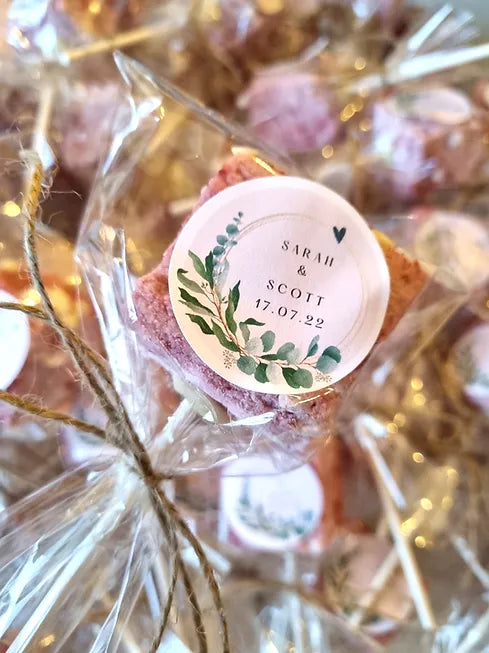 cellophane wrapped marshmallow lollie with sticker on, bride and grooms name on. held over table full of these same favours