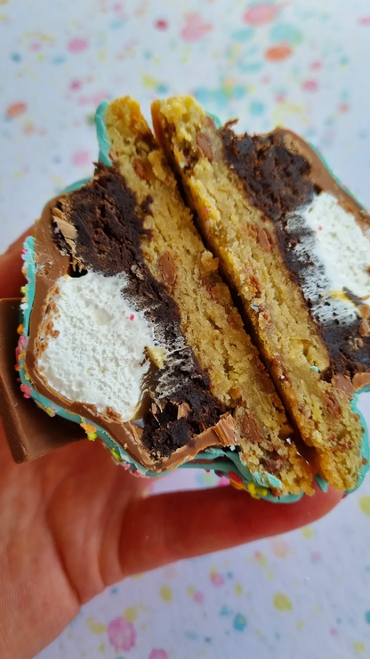 a cookie cut in half, shows the many layers of its inside. A layer of brownie, with a white fluffy marshmallow centre, topped with a chocolate top to seal it all in. This cookie is being held in tfhe palm of a hand above a lightly colourful background.