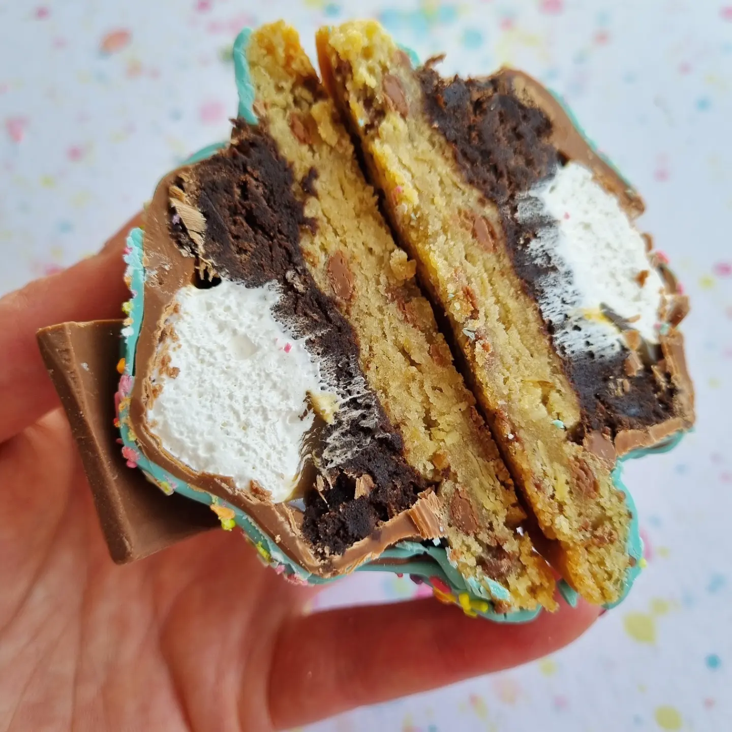a cookie cut in half, shows the many layers of its inside. A layer of brownie, with a white fluffy marshmallow centre, topped with a chocolate top to seal it all in. This cookie is being held in tfhe palm of a hand above a lightly colourful background.