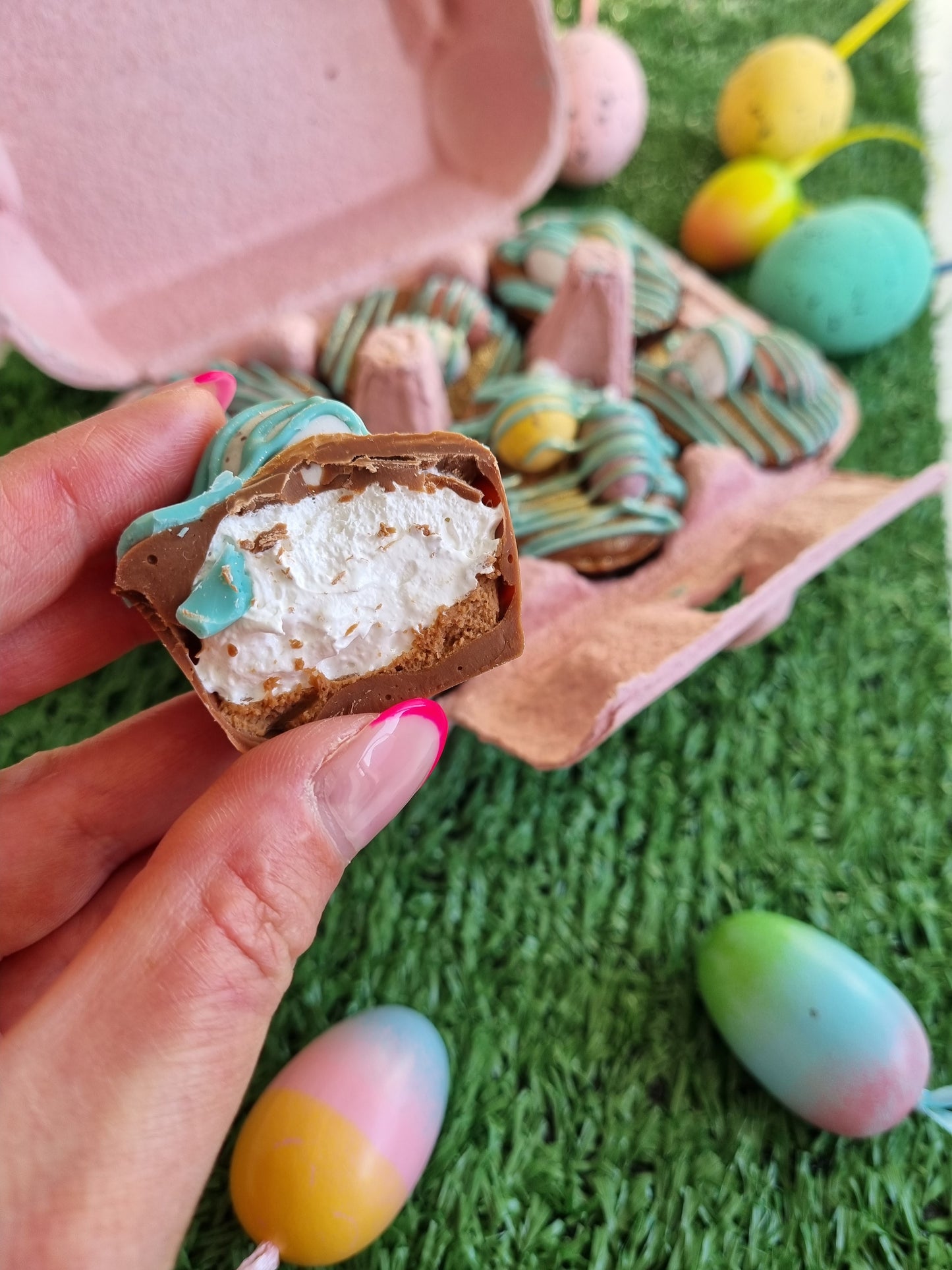 a hand is holding a little chocolate cup, there is a bite out of one and it is filled with fluffy white marshmallow. This is taken out of a pink egg box and inside the box is another 5 cups, each is drizzled in blue chocolate and topped with chocolate mini eggs. The box is sat on a fake green grass surface and their are coloured eggs around the egg box.