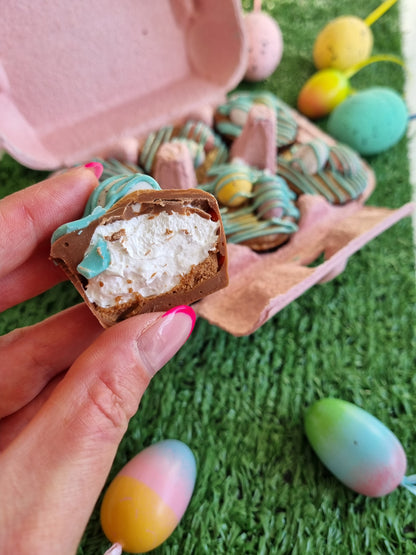 a hand is holding a little chocolate cup, there is a bite out of one and it is filled with fluffy white marshmallow. This is taken out of a pink egg box and inside the box is another 5 cups, each is drizzled in blue chocolate and topped with chocolate mini eggs. The box is sat on a fake green grass surface and their are coloured eggs around the egg box.