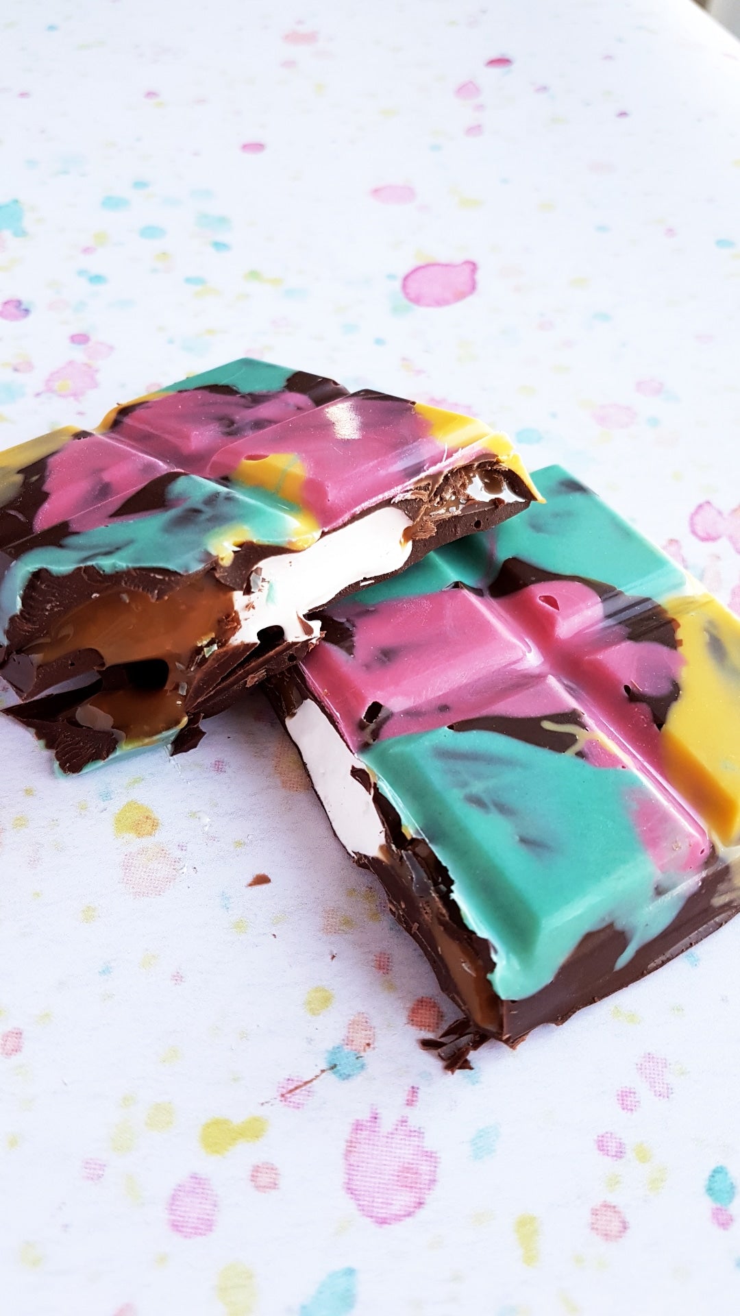 A Handmade Marshmallow Fluff Filled Chocolate bar, sits on top of a colourful speckled background. The chocolate bar is broken in half and the middle opf the chocolate bar which is a creamy white marshmallow fluff is oozing out of the middle. The chocolate bar is painted pink, green and yellow with colourful chocolate.