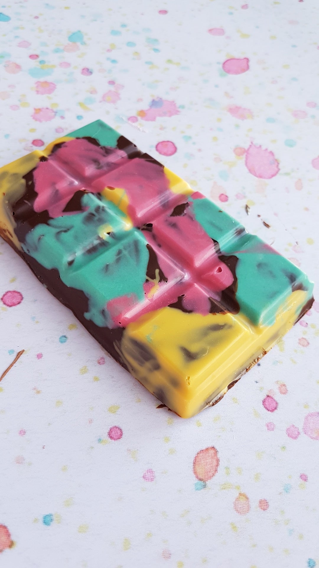 A Handmade Marshmallow Fluff Filled Chocolate bar, sits on top of a colourful speckled background. The chocolate bar is painted pink, green and yellow with colourful chocolate.