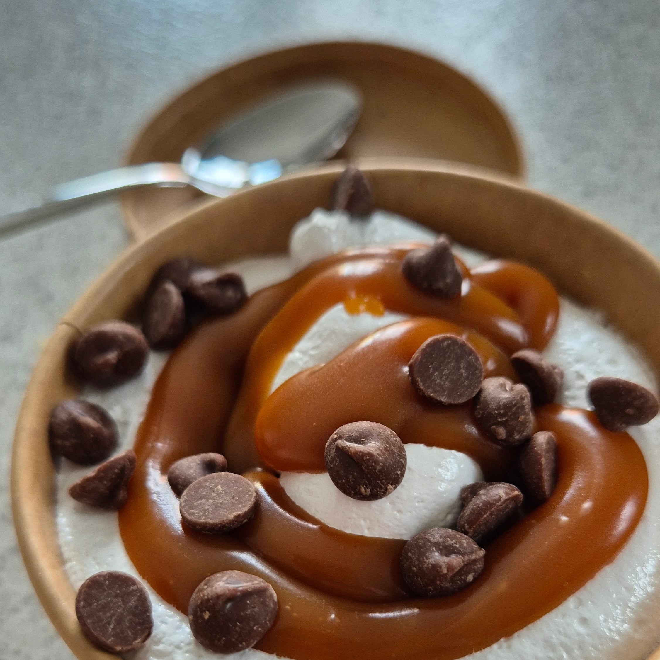 marshmallow fluff tub, with a layer of salted caramel on the top and chips of chocolate. tub has a spoon on its lid as its sat on top of table.