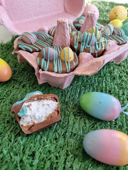 a hand is holding a little chocolate cup, there is a bite out of one and it is filled with fluffy white marshmallow. This is taken out of a pink egg box and inside the box is another 5 cups, each is drizzled in blue chocolate and topped with chocolate mini eggs. The box is sat on a fake green grass surface and their are coloured eggs around the egg box.