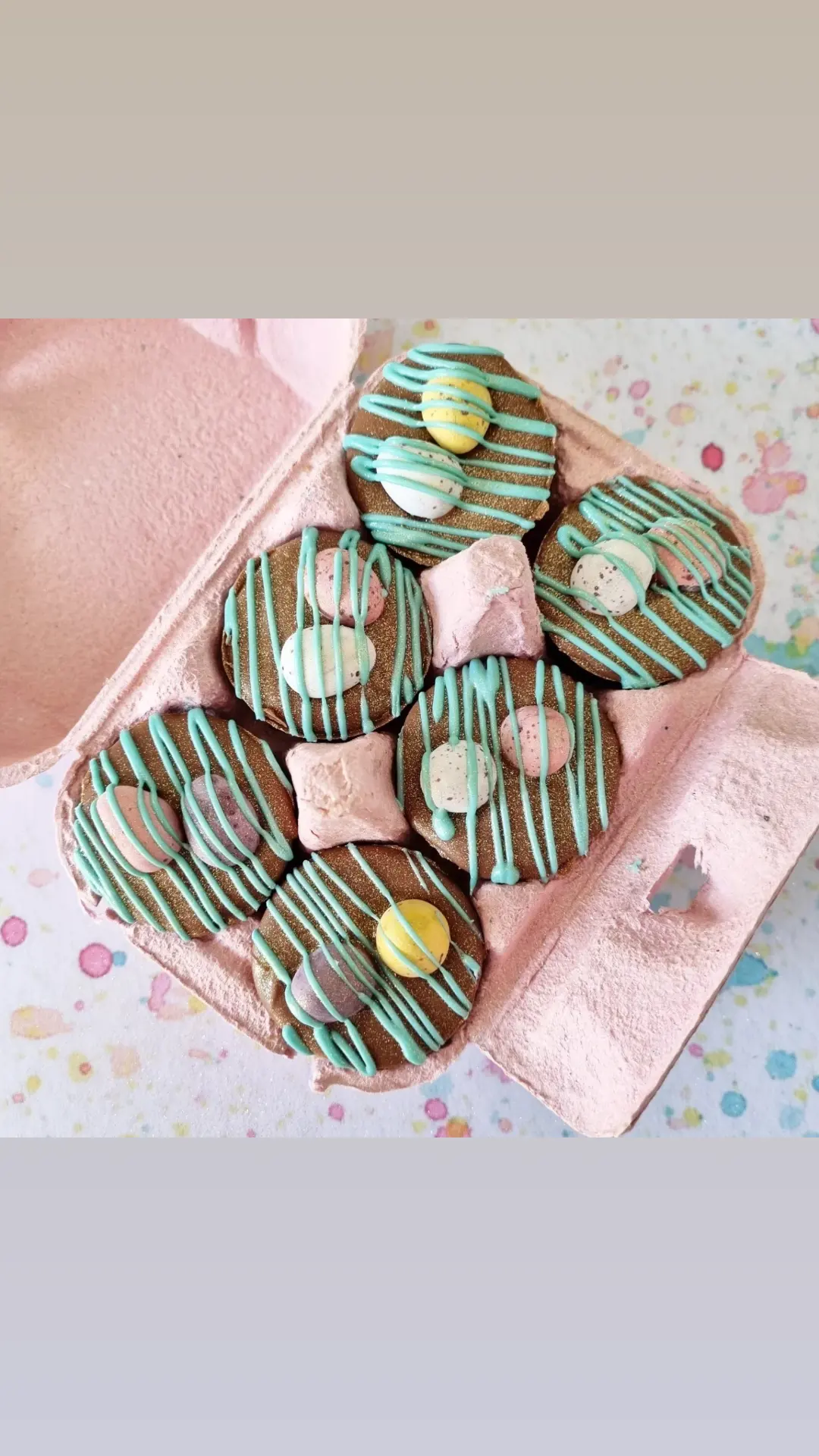 There are 6 chocolate mini cups in a  pink egg box . Each is drizzled in blue chocolate and topped with chocolate mini eggs. The box is sat on a light multi coloured surface.