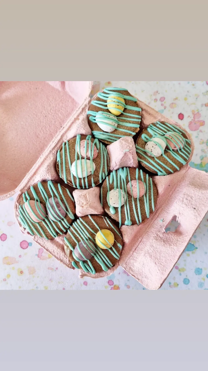 There are 6 chocolate mini cups in a  pink egg box . Each is drizzled in blue chocolate and topped with chocolate mini eggs. The box is sat on a light multi coloured surface.