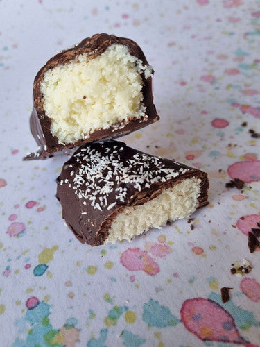 picture shows a handmade coconut bar, cut into tow pieces and stacked on top of each other. The inside of the bar is a white & creamy coconut filling, then dipped in chocolate and sprinkled with coconut. They are lay on a flat surface with multi-coloured droplets printed on the surface