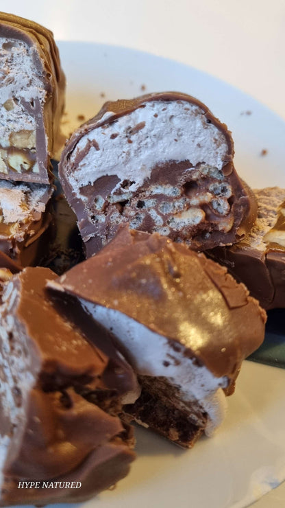 Handmade Chocolate Bar - Double Decker held by hand. layered chocolate crispies with a nougat and caramel top then all dipped in chocolate. all chopped up into bite size pieces on a plate. the plate is white in colour
