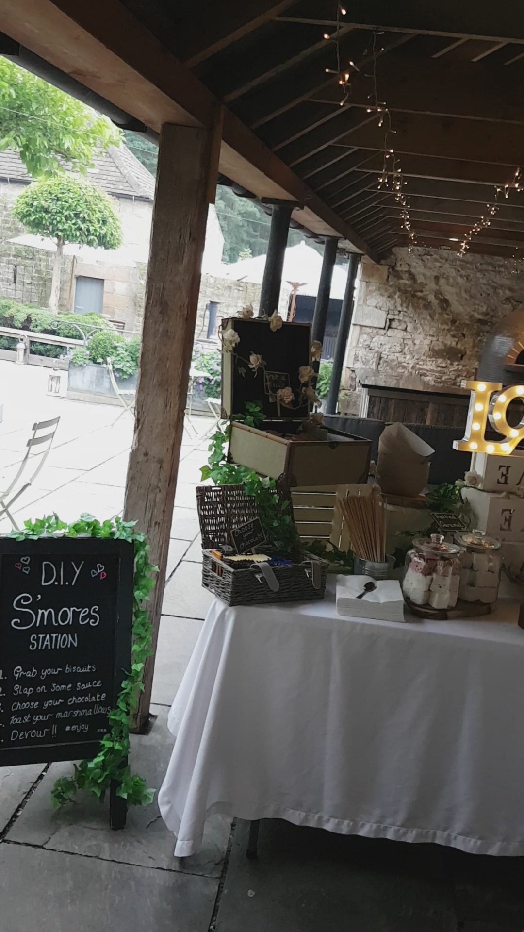 Load video: A full Smores set up station at Healey Barn wedding venue in Northumberland the North East Area, the table is set up underneath a wooden frame. The table holds boxes layered up &amp; jars top them filled with marshmallows, biscuits and chocolate.